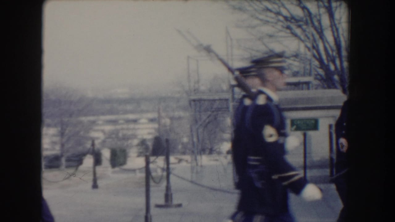 Uniformed honor guards march with rifles beside a stone railing and distant city view.