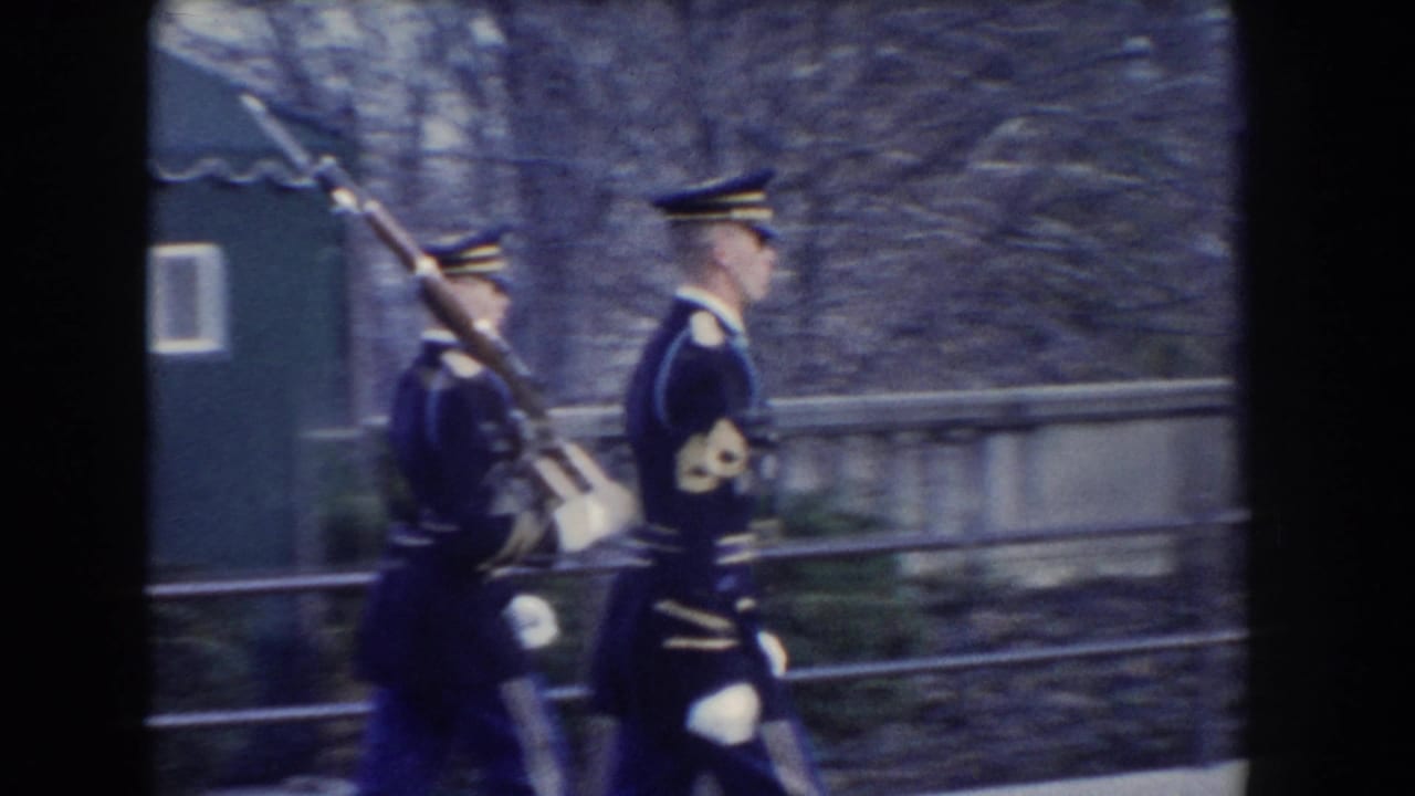 Two uniformed guards march past a stone wall and small blue structure.