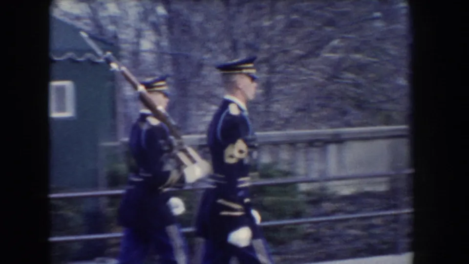 1960 Washington, DC frame of uniformed Air Force honor guards marching with rifles past a stone wall, bare trees, and a small structure.