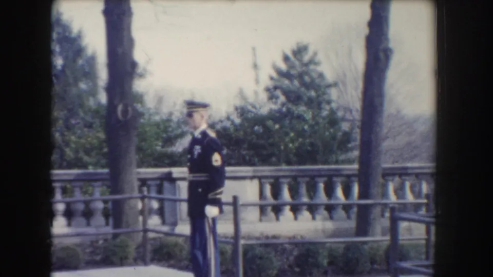 1960 Washington, DC home-movie frame showing a uniformed Air Force guard near a stone balustrade, walkway railing, shrubs, and bare trees.