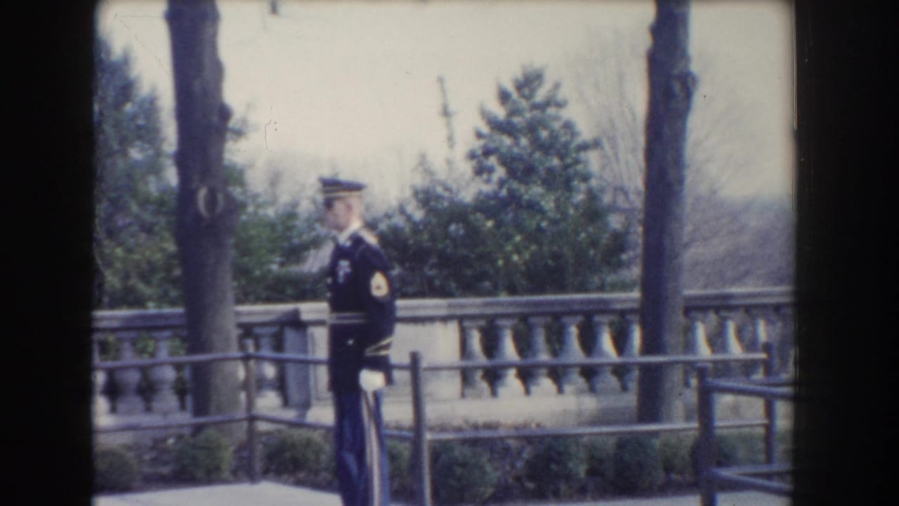 Uniformed guard in profile standing near a terrace railing with trees behind.