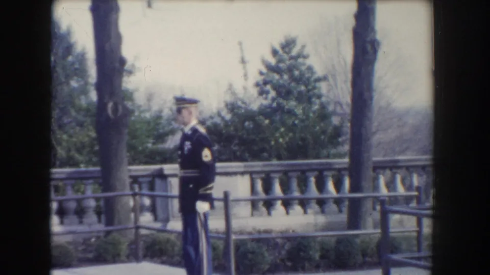 1960 Washington, DC frame of a formal uniformed guard standing in profile beside a terrace railing and stonework.