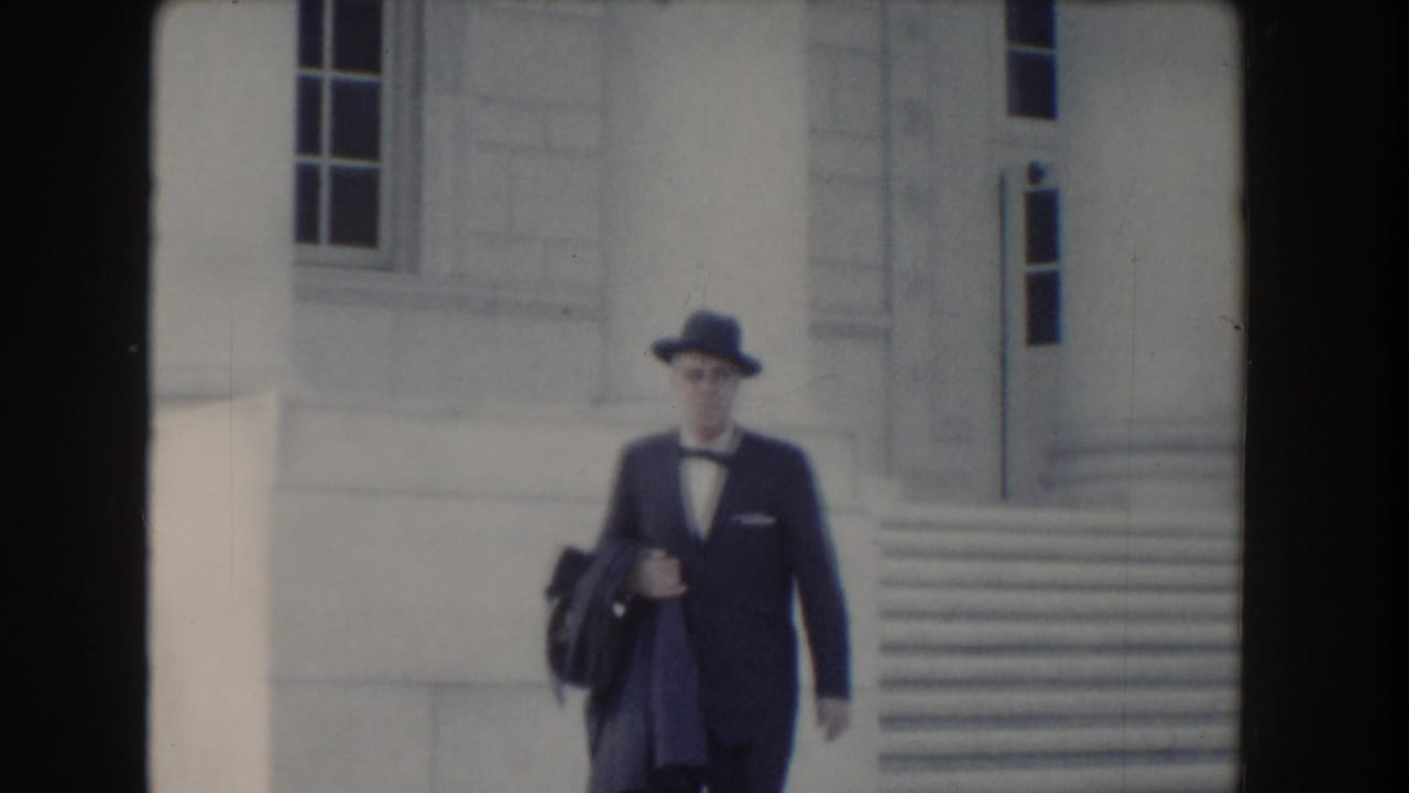 Man in a dark suit and fedora walks near stone steps outside a white building.