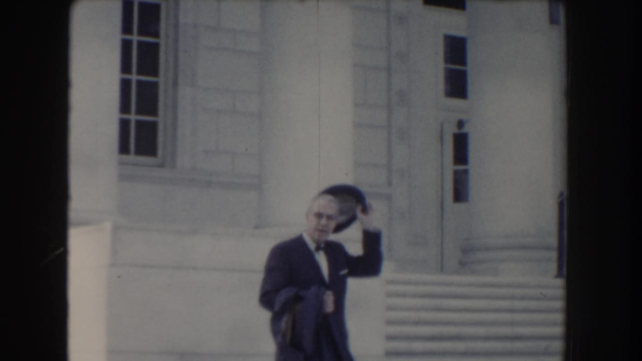 Suited man removes or touches his hat while walking in front of a white building.