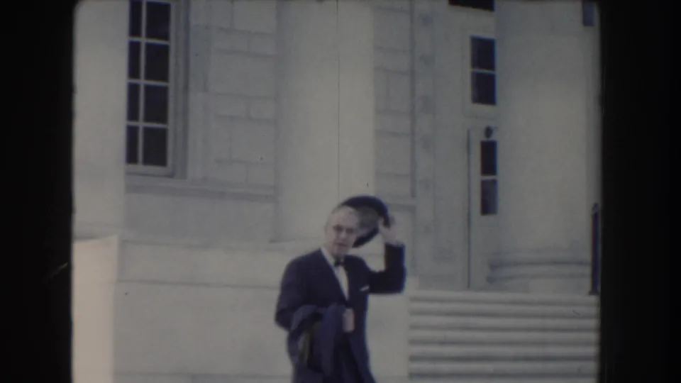 In Washington DC circa 1960, the suited man touches or removes his fedora as he walks past the white building facade.