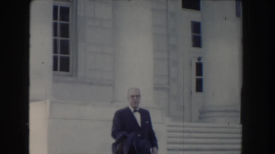 In Washington DC circa 1960, the man is closer to camera without his hat visible, carrying clothing over one arm near the steps.