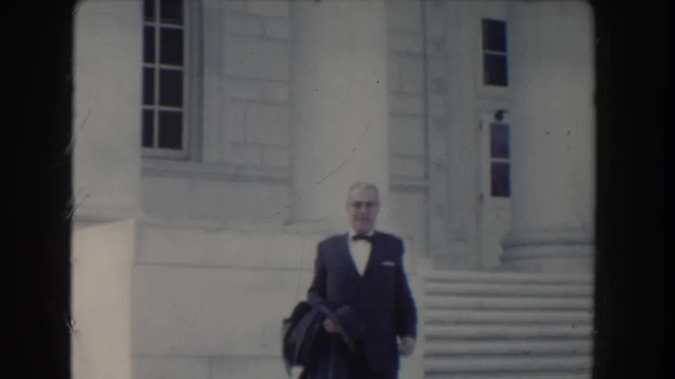In 1960 Washington, DC footage, a suited man with glasses carries outerwear while walking down steps beside large stone columns.