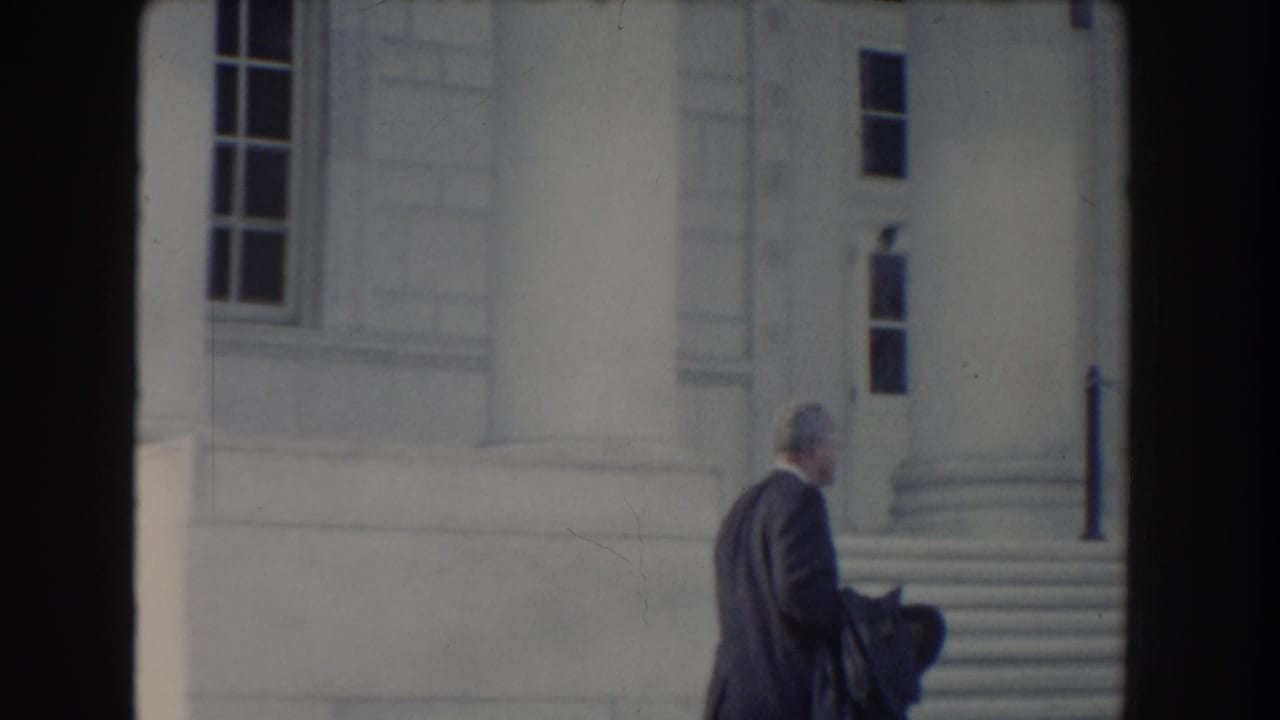 Suited man walks rightward past stone steps and columns, holding dark outerwear.