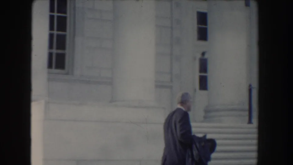 The man in a dark suit moves across the frame near broad steps and a classical columned exterior in Washington, DC.
