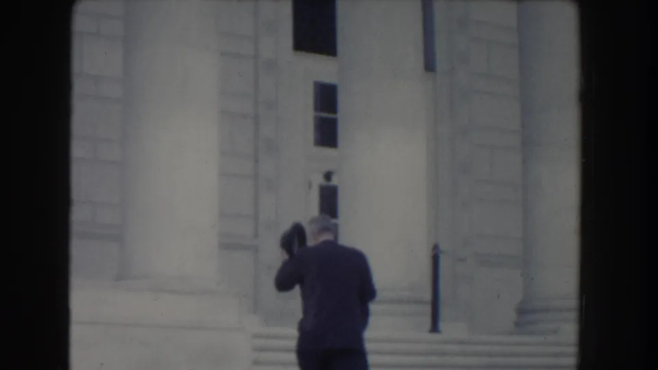 The suited man lifts an arm near the stone steps, with a large column, tall windows, and pale masonry visible behind him.