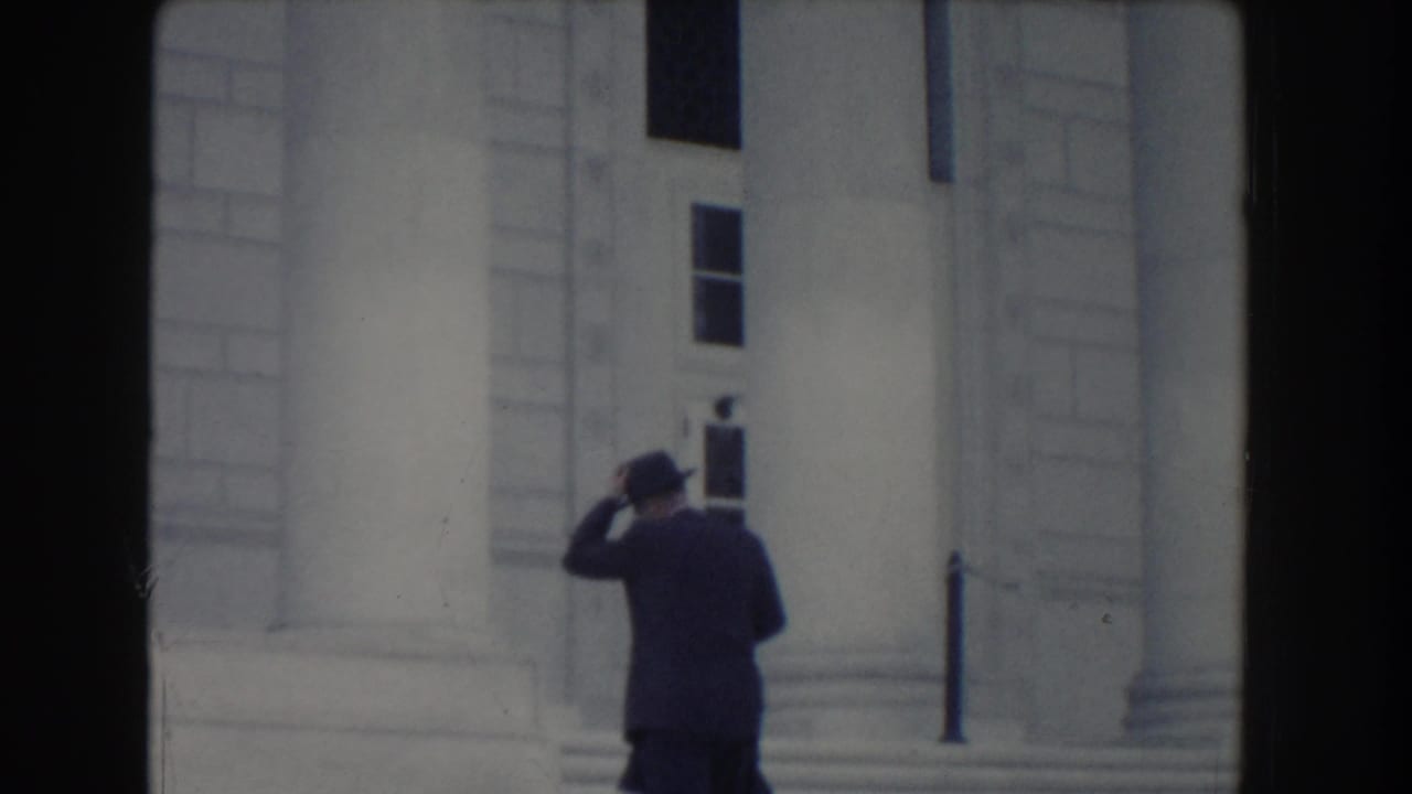 Man in a dark suit and fedora walking near stone steps and columns.