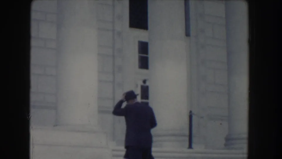 1960 Washington, DC home-movie frame showing a suited man in a fedora near the steps of a neoclassical building.