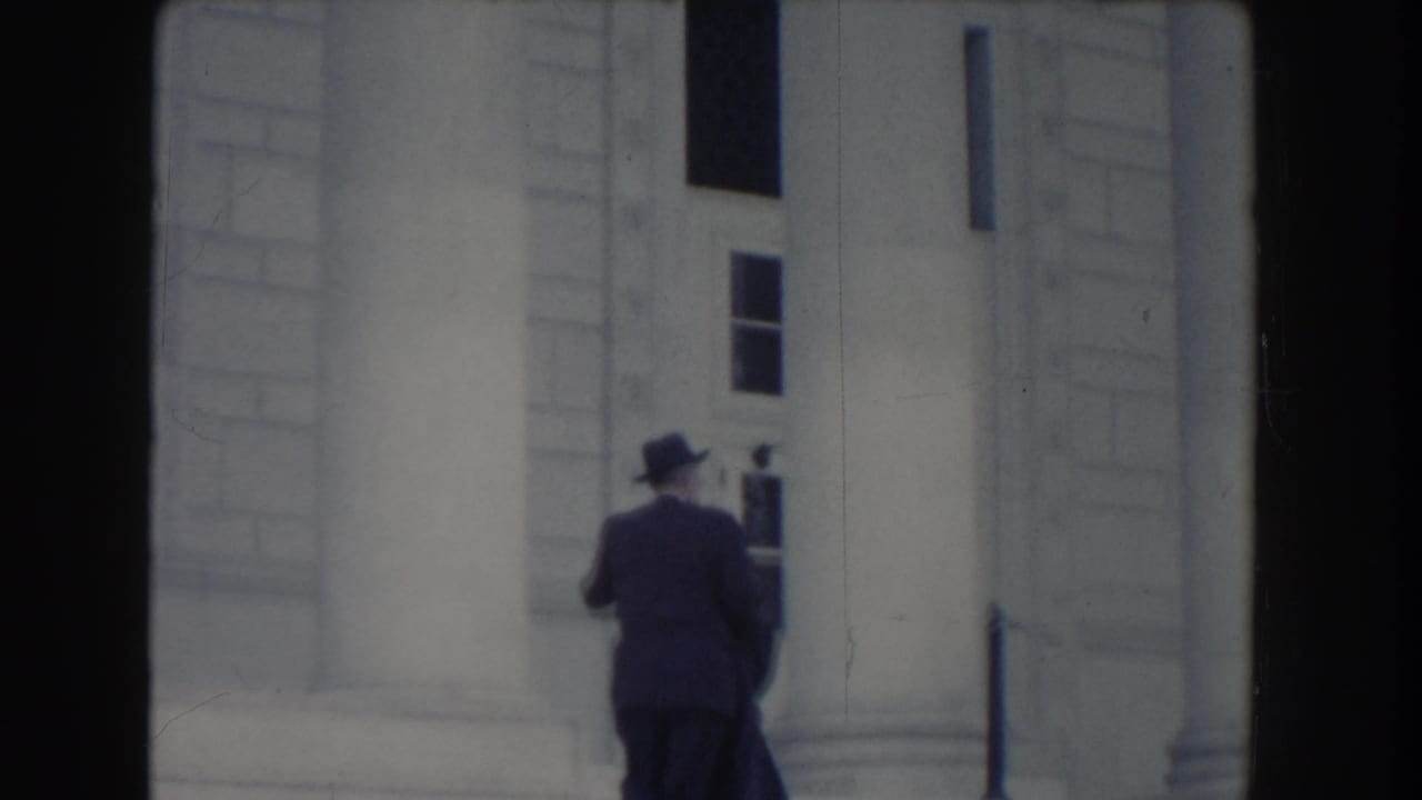 Suited man in a hat walking past a columned stone facade.