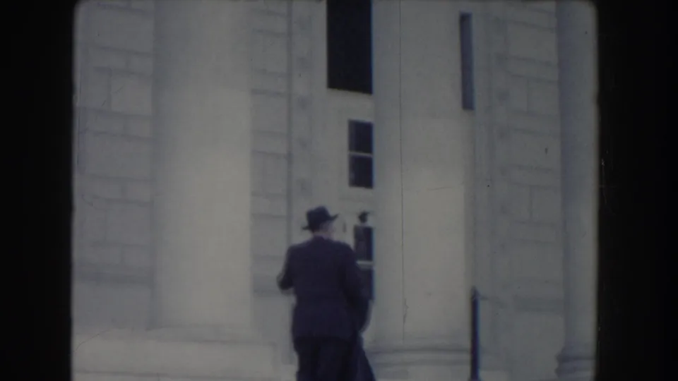 A dark-suited man in a fedora continues along a pale neoclassical building exterior in Washington, DC archival footage.