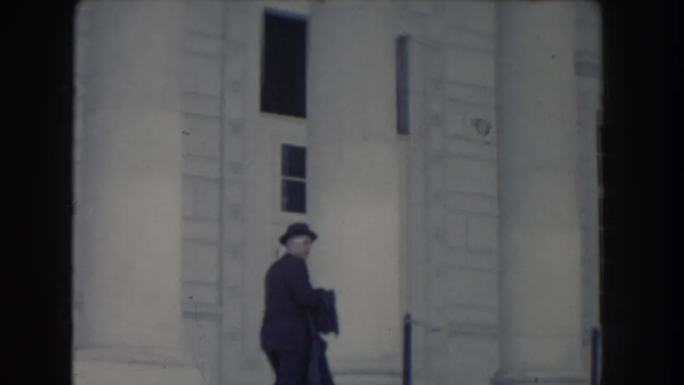 The fedora-wearing man appears to turn or glance back near the steps of a Washington, DC neoclassical building.