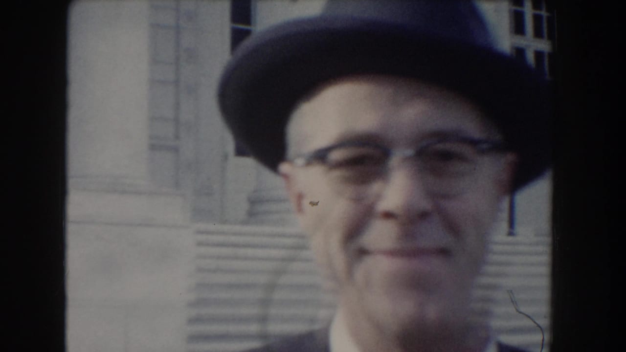 Man in a fedora and glasses smiles near building steps.