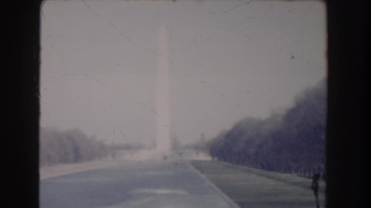 Distant 1960 home-movie frame of the Washington Monument and National Mall paths under a pale sky.