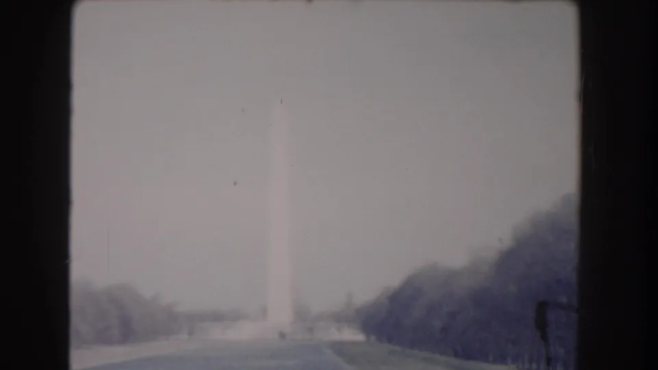 1960 8mm frame showing the Washington Monument in haze above lawns, walkways, and dark trees on the National Mall.