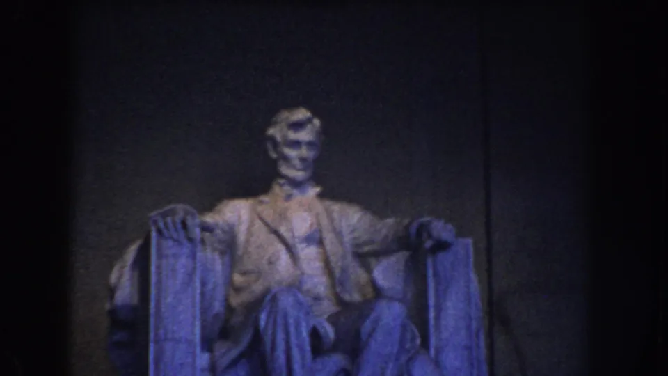 1960 Washington DC home-movie frame showing the seated Lincoln statue with knees, hands, and chair arms visible in low light.