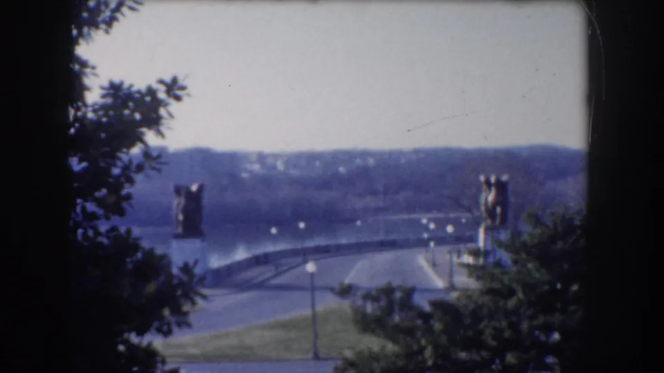 1960 Washington, DC footage frames a bridge and its statues through dark foreground foliage, with open land and distant terrain behind.