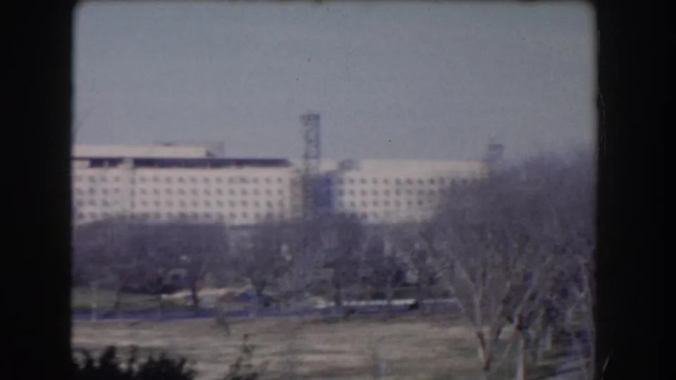 1960s Washington, DC home-movie frame showing a distant federal-style building behind bare trees and parkland.