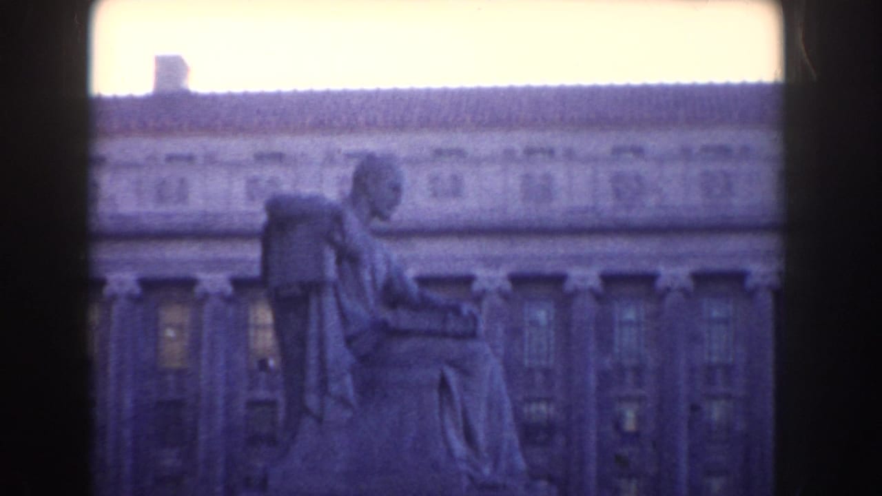 Seated stone statue in front of a large neoclassical building, seen through dark frame edges.