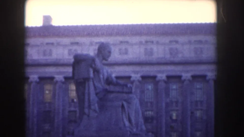 1960 Washington, DC home-movie frame showing a seated robed statue before a grand neoclassical facade, partially blocked by dark framing.