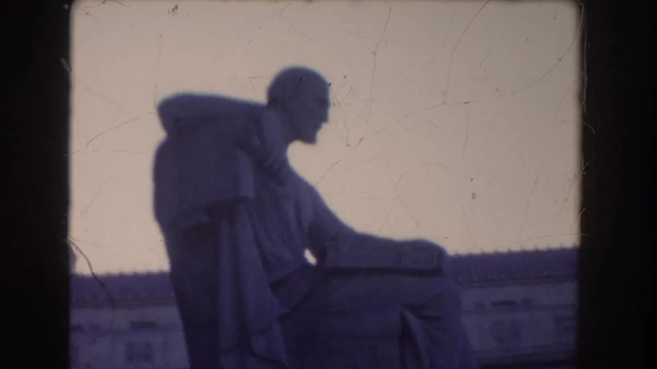 1960 Washington, DC home-movie still showing the seated robed statue in soft focus with a low neoclassical roofline behind it.