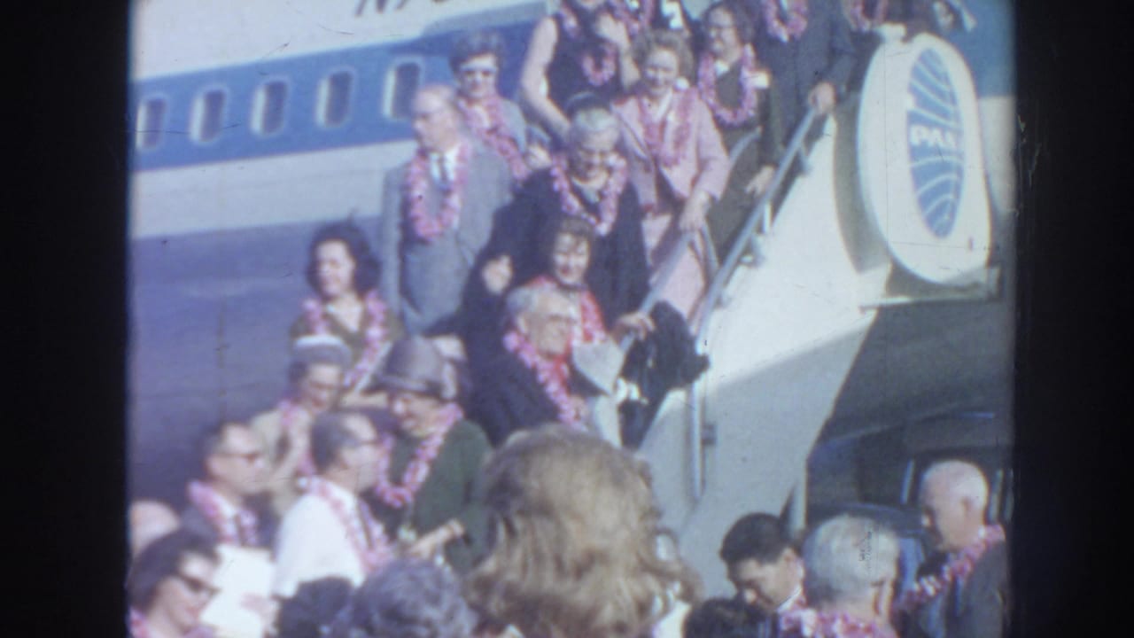 Passengers wearing leis descend stairs from a Pan Am aircraft as a crowd watches below.