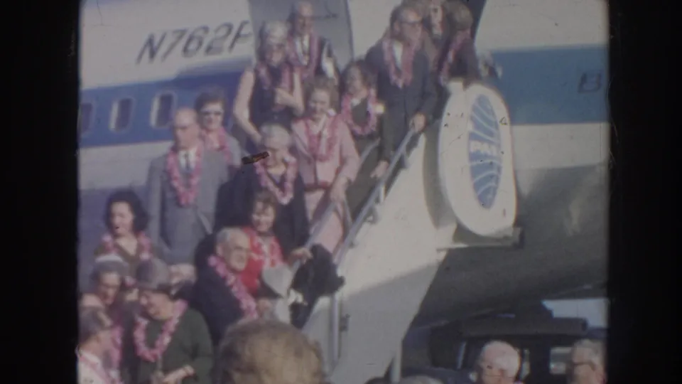 Lei-wearing travelers descend from a Pan Am aircraft in Hawaii, with faces visible and a dense arrival crowd below the stairs.