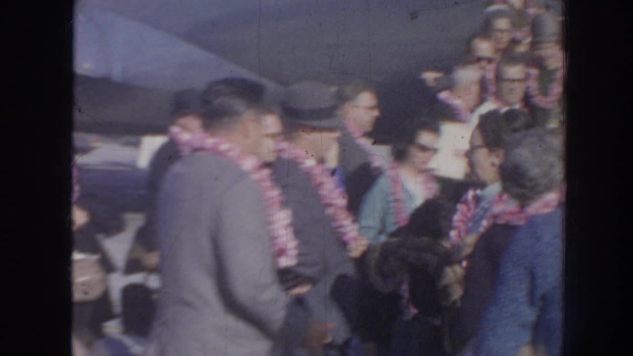 1961 Hawaii home-movie frame showing travelers in leis, suits, hats, and sunglasses gathered closely in an arrival crowd.