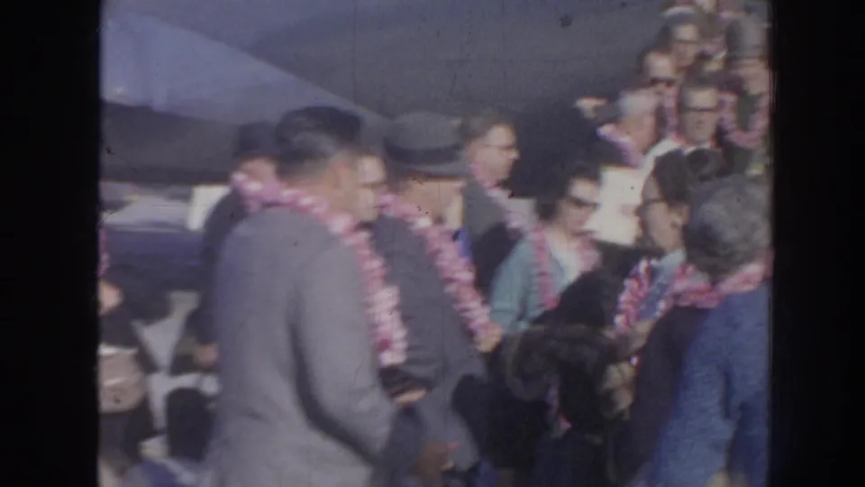 1961 Hawaii home-movie frame showing travelers in leis, suits, hats, and sunglasses gathered closely in an arrival crowd.