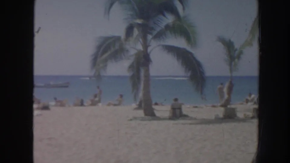 1961 Hawaii home-movie frame showing palm trees, beachgoers on sand, blue ocean, and small boats offshore.