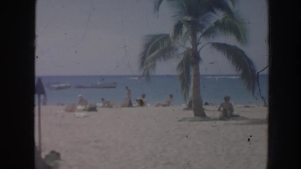 1961 Hawaii beach frame with a leaning palm, people seated on sand, ocean water, and small boats visible offshore.