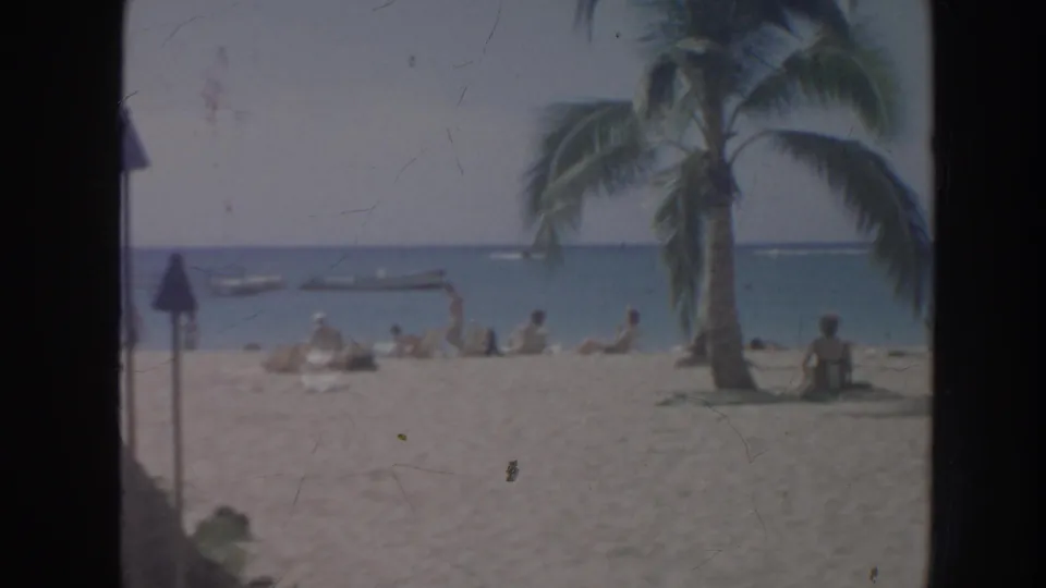 1961 Hawaii home-movie frame showing beachgoers on a sandy shore with palms, ocean, and small boats in the distance.