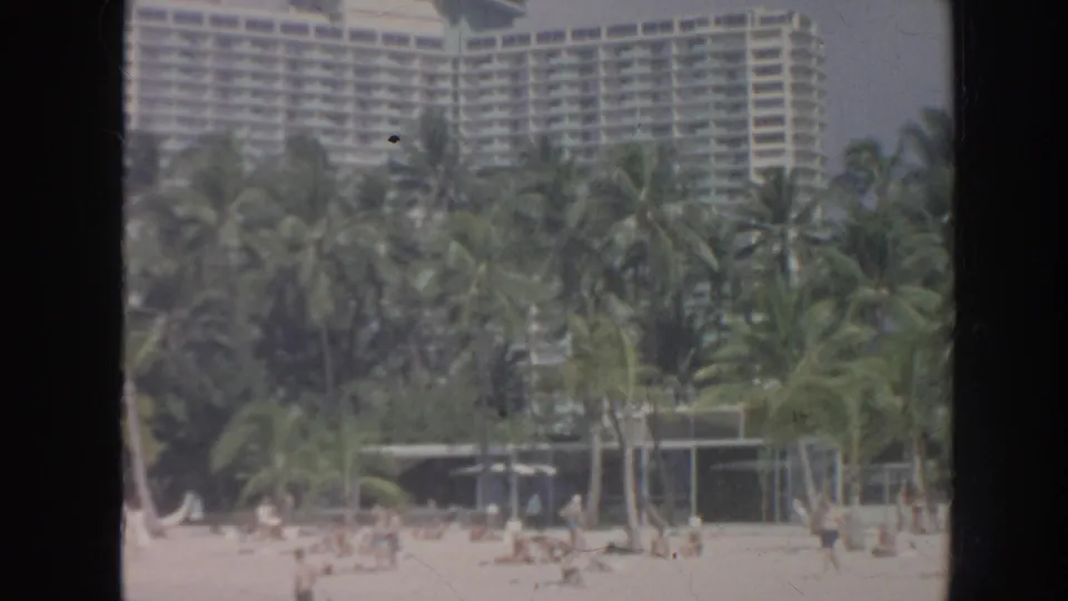 1961 Hawaii frame showing a large multi-story hotel behind palms, with beachgoers scattered across the sand in front.