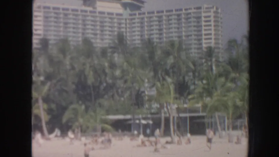 1961 Hawaii home-movie view of a large hotel beyond palms, with people on the beach and soft faded color.