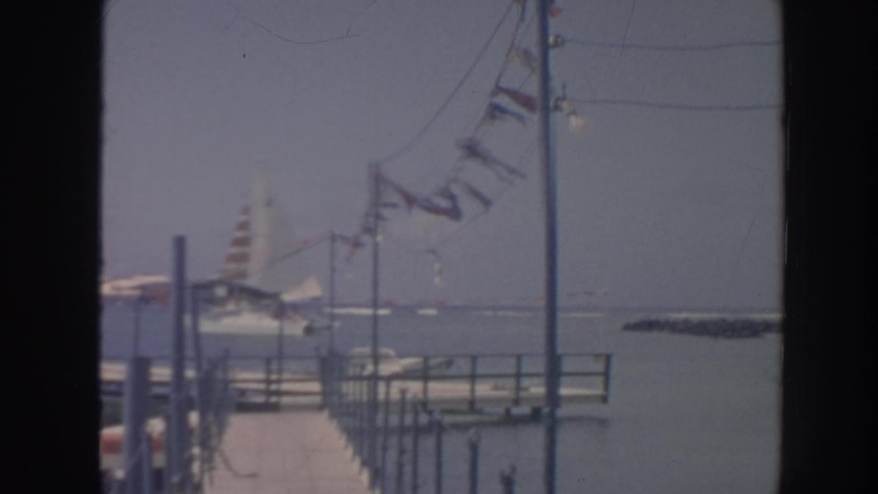 Marina pier with fluttering flags, a sailboat, and sea beyond.