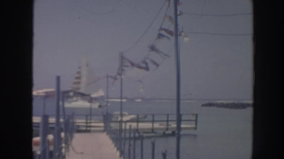 1961 Hawaii home-movie frame showing a waterfront pier, flags in strong wind, a sailboat, and open water.