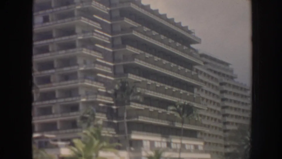 1961 Hawaii home-movie frame of a large tiered hotel facade with balconies and palms seen through a dark frame.