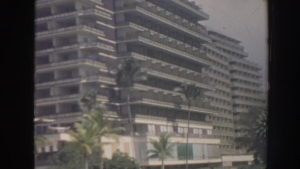 1961 Hawaii footage frame showing a mid-century hotel exterior with stacked balconies, palms, and soft 8mm color.