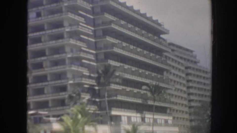 1961 Hawaii home-movie frame of a resort hotel facade, palm trees, and a dark window frame cutting through the view.