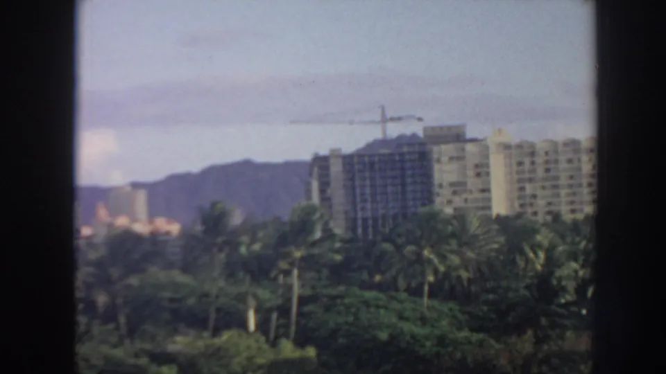 1961 Hawaii coastal development scene with high-rise buildings, a crane, palms, and distant mountains beyond the shoreline.