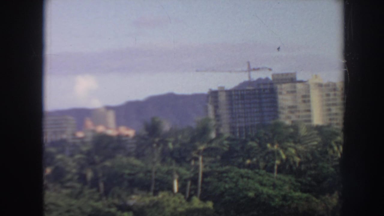 1961 Hawaii home-movie view across palms toward high-rise buildings under construction and hazy hills.