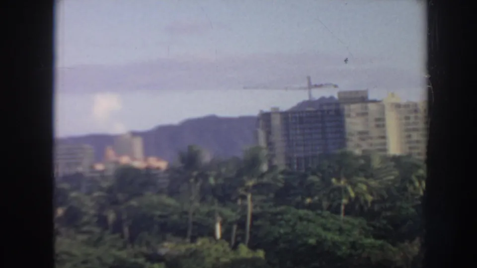 1961 Hawaii home-movie view across palms toward high-rise buildings under construction and hazy hills.