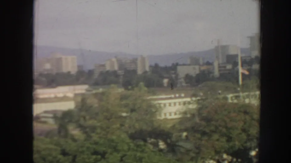 1961 Hawaii home-movie frame with soft focus on trees, low buildings, high-rises, and hazy hillside background.