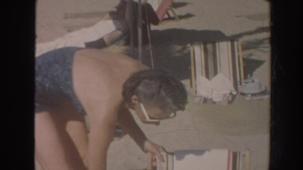 In 1961 Hawaii home-movie footage, a woman in a blue swimsuit and white sunglasses bends near a striped beach chair.