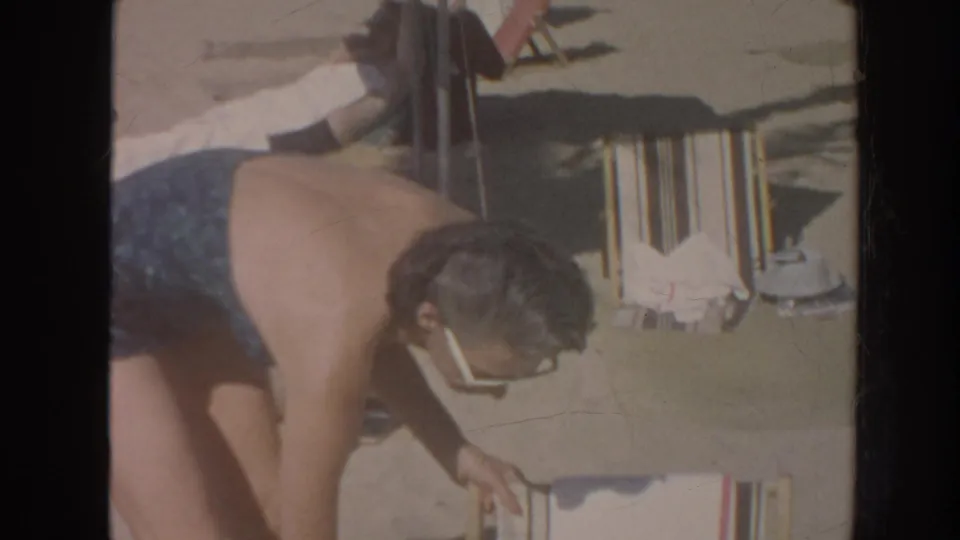 In 1961 Hawaii home-movie footage, a woman in a blue swimsuit and white sunglasses bends near a striped beach chair.