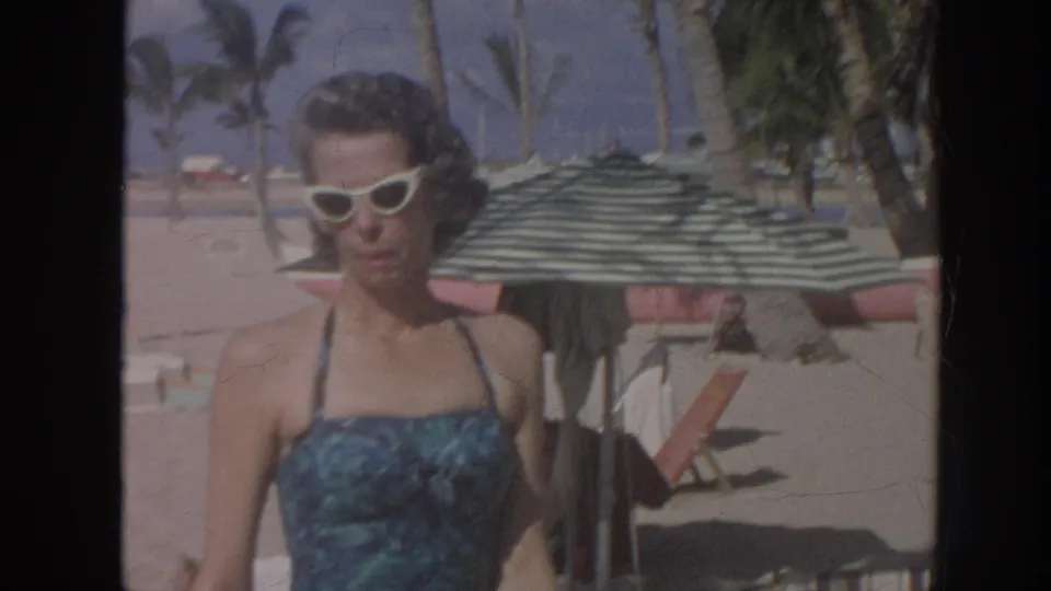 A woman in a blue patterned swimsuit and white cat-eye sunglasses faces the camera on a Hawaii beach with palms behind her.