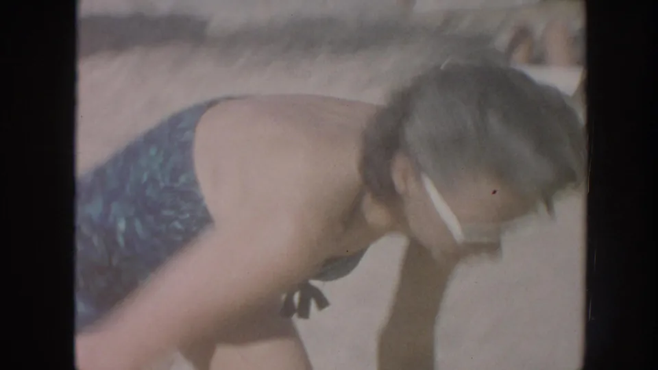 A close handheld frame shows the woman bending forward in white sunglasses and a blue swimsuit on the sandy beach.